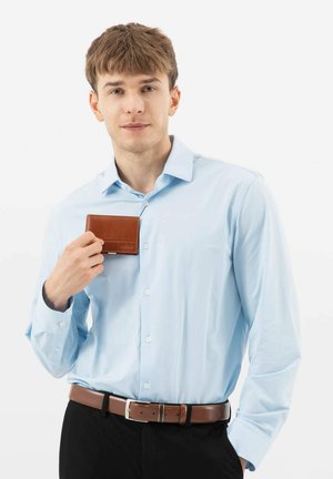 Lichtblauw overhemd met lange mouwen, gecombineerd met een bruine leren portemonnee die voor zich wordt gehouden. Zwarte broek en een bruine riem accentueren de outfit.