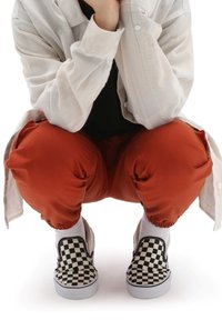 Scarpe slip-on a scacchi in bianco e nero su calze bianche, indossate con pantaloni color ruggine e una camicia a maniche lunghe di colore chiaro.