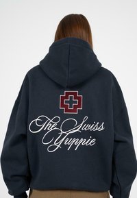 Granatowa, oversize bluza z kapturem z białym napisem "The Swiss Guppie" oraz bordowym logo krzyża szwajcarskiego na plecach. Materiał bawełniany, luźny krój.