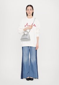Sweatshirt blanc avec texte rouge, associé à un jean en denim à jambes larges avec des panneaux latéraux foncés, et tenant un petit sac à main en argent.