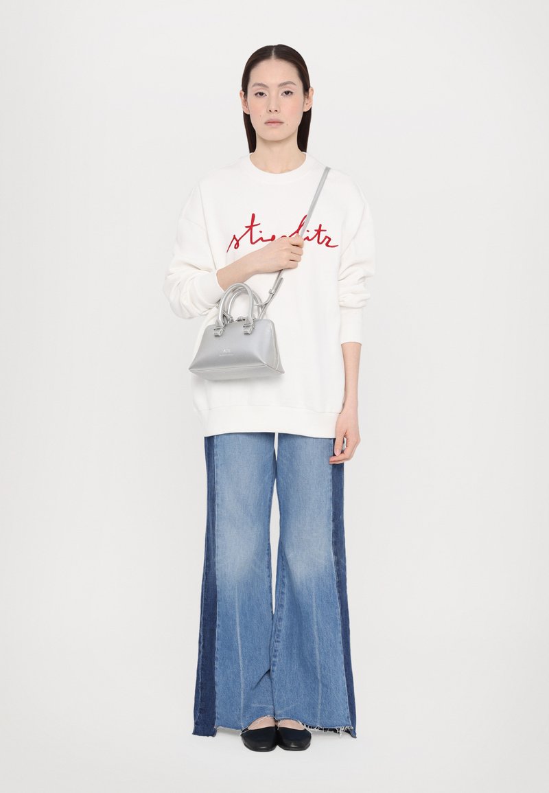 Sweatshirt blanc avec texte rouge, associé à un jean en denim à jambes larges avec des panneaux latéraux foncés, et tenant un petit sac à main en argent.
