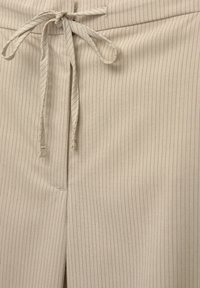 Tissu beige avec de fines rayures verticales noires, doté d'une taille à cordon noué et d'une couture de braguette frontale dissimulée.