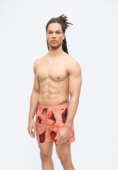 Homme musclé aux dreadlocks debout, portant un short de bain orange avec des motifs abstraits noirs et rouges sur un fond blanc uni.