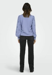 Kvinna som står med ryggen mot kameran, klädd i en lila långärmad tröja med brodyrdetaljer på ärmarna och svarta raka jeans.