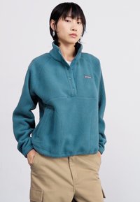 Pull en polaire teal avec un col haut, fermeture à boutons pression et poche avant. Matériau texturé et coupe décontractée.