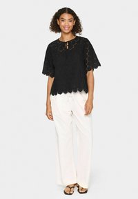 Top in pizzo nero con maniche corte, orlo scalloppato e dettaglio con apertura a chiave e nodo. Indossato con pantaloni bianchi a gamba larga e sandali neri.