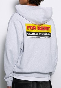 Szara bluza z dużym, wyraźnym napisem "DO WYNAJĘCIA" na plecach w czerwonym i żółtym kolorze, zawierająca dane kontaktowe w czerni i bieli. Materiał bawełniany.