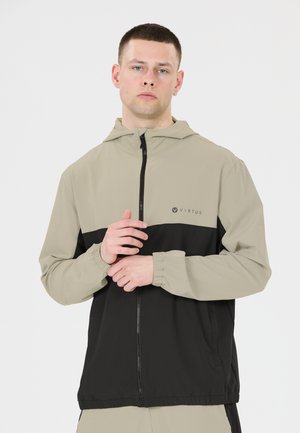 Mand iført en to-farvet beige og sort lynlåstrøje med hætte, stående mod en ensfarvet hvid baggrund.