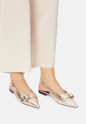 Metaliczne złote buty na obcasie z odkrytą piętą, z ostrym noskiem, ozdobione klamrą i teksturowanym wykończeniem, zestawione z jasnymi, szerokimi spodniami.