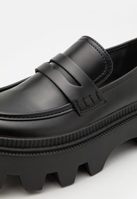 Mocassin en cuir noir avec semelle épaisse à crans et détail de bride sur le dessus lisse.