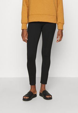 Leggings noirs au design ajusté, avec une texture lisse et sans coutures visibles. Portés avec un sweat-shirt moutarde et des sandales à enfiler noires.