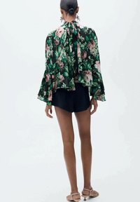 Blouse florale en tissu vert foncé, avec des imprimés de fleurs roses et blanches. Dotée de manches longues évasées et d'un dos à ouverture en forme de trou de serrure. Associée à un short noir.