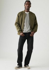 Man in olive groene bomberjack, wit en zwart gestreept shirt, zwarte jeans en zwarte sneakers, staande tegen een effen lichte achtergrond.