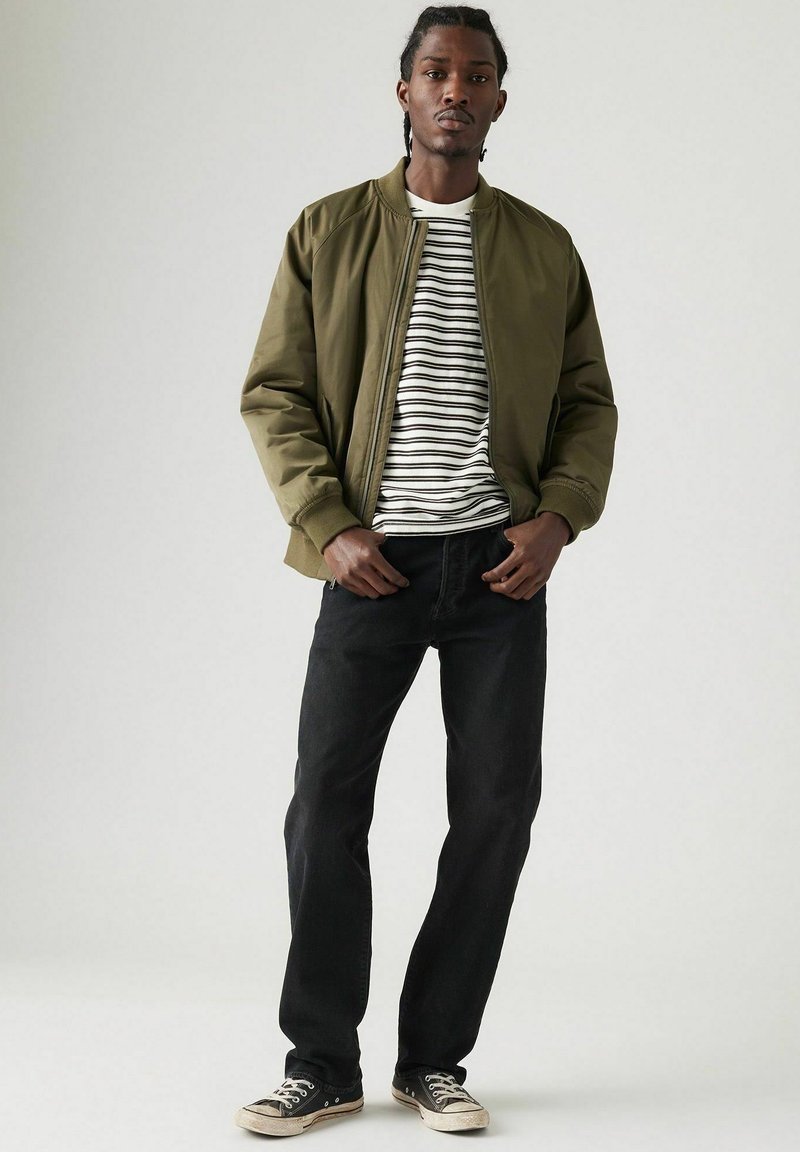 Man in olive groene bomberjack, wit en zwart gestreept shirt, zwarte jeans en zwarte sneakers, staande tegen een effen lichte achtergrond.