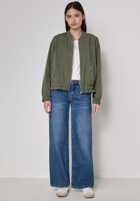 Giacca bomber verde con colletto a costine, chiusura con zip e tasche laterali; abbinata a jeans blue a gamba larga e sneakers bianche. Tessuto morbido.