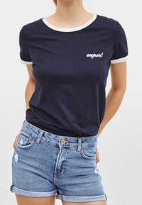 Camiseta de la marina con cuello y puños blancos, con la palabra "¡super!" en letras blancas. Combinada con pantalones cortos de mezclilla desgastados.