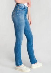 Jeans bleus slim dans un délavé, dotés de cinq poches, d'une fermeture à glissière et de détails en métal doré. Portés avec des baskets blanches.