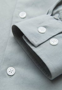 Poignet de chemise à manches longues gris clair avec boutons blancs à quatre trous et couture visible sur le tissu.