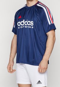 Man som bär en marinblå Adidas sporttröja med röda och vita ränder och vita Adidas shorts med logotyp på vänster ben.