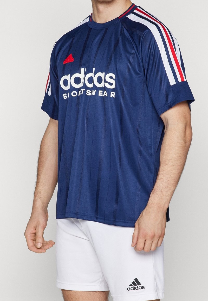 Man som bär en marinblå Adidas sporttröja med röda och vita ränder och vita Adidas shorts med logotyp på vänster ben.