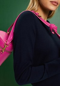 Sac à main en cuir rose de forme arrondie, avec des accessoires dorés et une longue bandoulière, porté sur un pull marine devant un fond vert.