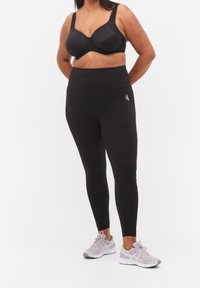 Czarny sportowy stanik i legginsy z wysokim stanem, wykonane z elastycznego materiału. Tenisówki są jasnoszare, z oddychającej siateczki.