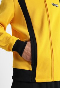 Gul textiljacka med svarta detaljer, dragkedja fram, ribbade ärmslut och sidofickor. Strukturerad tyg ger hållbarhet.