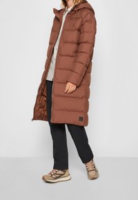 Brun puffjacka med huva, med en kviltad design, framzip och sidofickor. Matchas med mörka byxor och bruna sneakers.