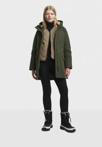 Mujer vestida con ropa de invierno en capas, incluyendo un abrigo verde, un chaleco acolchado color beige, leggings negros y botas negras de invierno, de pie contra un fondo sencillo.