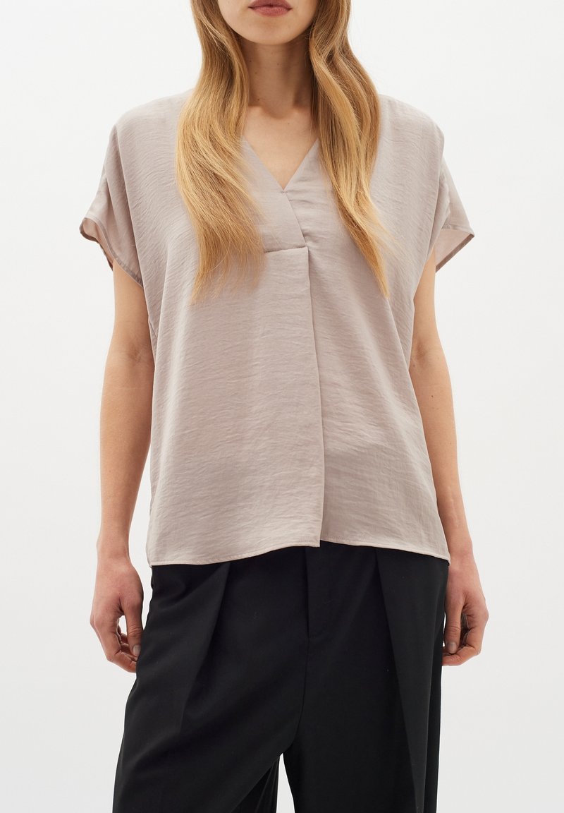 Femme portant un chemisier ample beige clair à manches courtes et col en V, et un pantalon noir taille haute, debout devant un fond blanc.