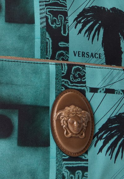 Emblème tête de Méduse marron sur tissu bleu sarcelle avec motifs noirs, palmiers et le mot Versace.