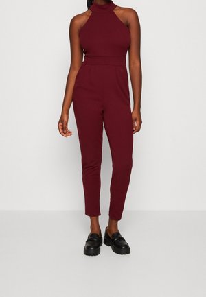 Bordowy kombinezon z wysokim dekoltem, bez rękawów, ze zwężanymi nogawkami i dopasowaną talią. Noszony z czarnymi wsuwanymi butami.