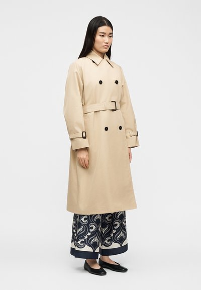 Γυναίκα που φοράει μπεζ διπλής σειράς καμπαρντίνα με ζώνη, φ αρδιά παντελόνια με σχέδιο και μαύρες φλατ, στέκεται μπροστά σε απλό λευκό φόντο.