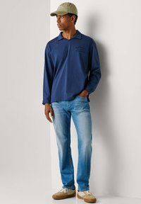 Man met een olijfgroene baseballpet, donkerblauw shirt met lange mouwen, lichtblauwe spijkerbroek en beige sneakers, staand tegen een witte muur.