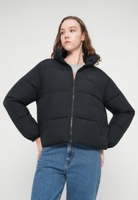 Μαύρο μπουφάν puffer με ψηλό γιακά, κλείσιμο με φερμουάρ και πλαϊνές τσέπες. Διαθέτει οριζόντια καπιτονέ πάνελ και λογότυπο στο στήθος.