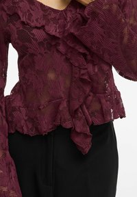 Blouse en dentelle bordeaux avec un décolleté et des manches volants, présentant un motif floral et une texture transparente, associée à un pantalon noir.