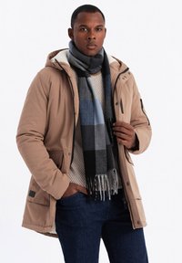 Giacca isolata marrone con cappuccio, indossata sopra un maglione leggero. Jeans scuri e una sciarpa a quadri multicolore completano l'outfit.