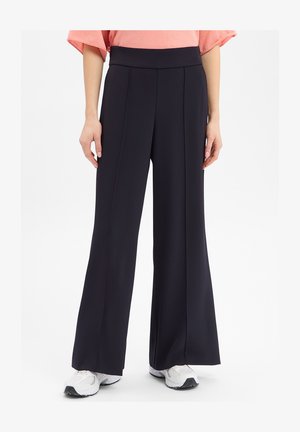 Frau trägt schwarze Hosen mit weitem Bein, weiße Sneakers und ein korallenfarbenes Kurzarmoberteil, steht vor einem schlichten weißen Hintergrund.