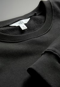 Svart sweatshirt i bomull med ribbad rund hals och mudd. Inkluderar ett tygmärke med varumärkes- och storleksinformation.
