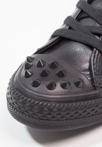 Black sneakers feature a glossy leather upper with a round toe capped in textured rubber and adorned with flat black studs.