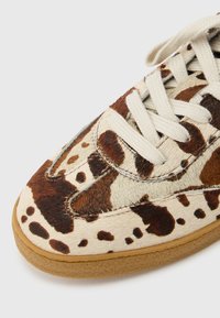 Αθλητικό παπούτσι με ανοιχτό καφέ λαστιχένια σόλα και επάνω μέρος με σχέδιο από δέρμα αγελάδας σε κρεμ, καφέ και σκού ρο καφέ αποχρώσεις, με λευκά επίπεδα κορδόνια.