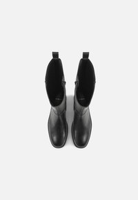Botas de tornozelo em couro preto com textura lisa, apresentando um formato alongado, painéis laterais elásticos e abas para facilitar o calçar.
