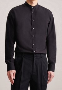 Camicia nera a maniche lunghe con collo alla coreana, taglio dritto, con una lieve lucentezza e bottoni davanti, abbinata a pantaloni neri.