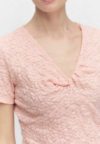 Lyserød kjole med et struktureret blondermønster, V-hals, korte ærmer og indsnøret detalje foran.