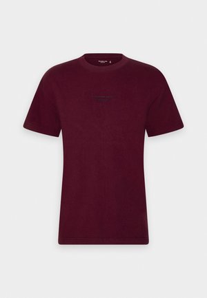 T-shirt in cotone bordeaux con scollo rotondo e maniche corte. Presenta un piccolo logo stampato di colore verde scuro sul davanti.