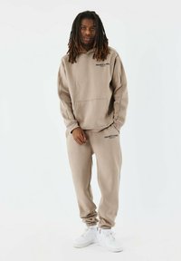 Conjunto de sudadera y pantalones de chándal beige, que incluye un bolsillo frontal, logotipo de texto "PROJECT X PARIS" y puños acanalados, combinado con zapatillas blancas.