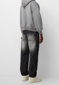 Persoon die een grijze hoodie, zwarte verweerde spijkerbroek met grote achterzakken en witte sneakers draagt, staand tegen een effen lichte achtergrond.