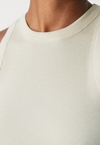 Top a canottiera a righe di colore crema chiaro, caratterizzato da un collo alto, spalline larghe e una texture liscia. La vista ravvicinata mette in risalto i dettagli del tessuto.