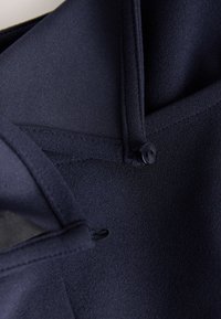 Gros plan sur un tissu bleu marine avec un petit bouton noir fixant une sangle bouclée. Le tissu a une finition texturée et lisse.