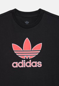 Melna kokvilnas T-kreklu ar sarkano un baltu Adidas logotipu, kas satur trīs lapu dizainu un trīs horizontālas svītras zem zīmola nosaukuma.
